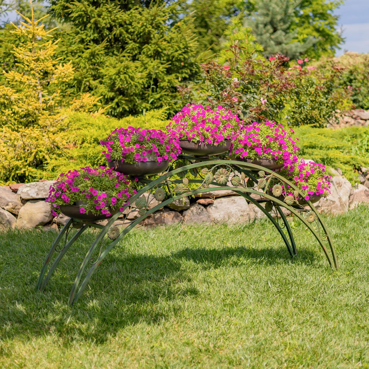 Arched green metal plant stand on a lawn, holding flower-filled bowls among garden greenery and rocks.