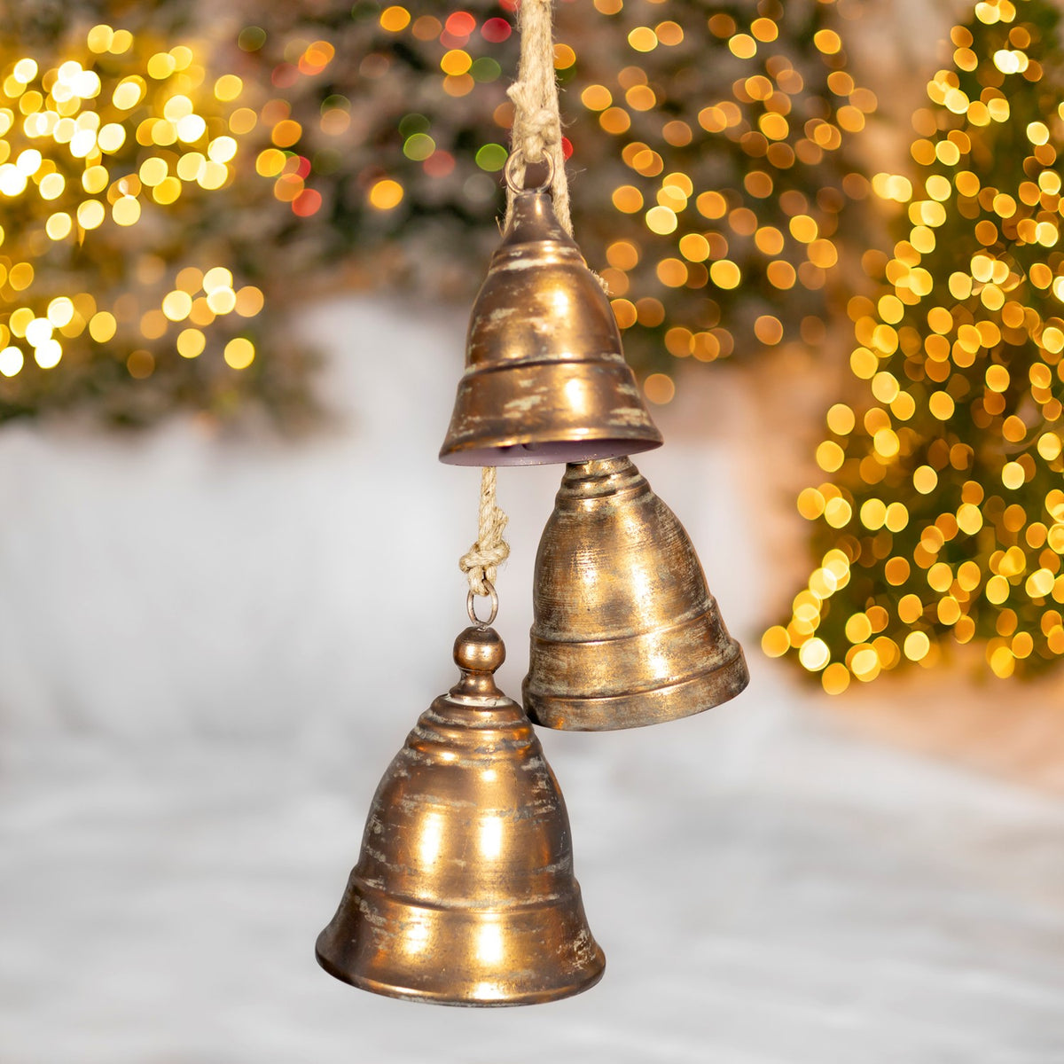 Three differently sized bronze-gold bells hang from rough cords against warm twinkling lights.