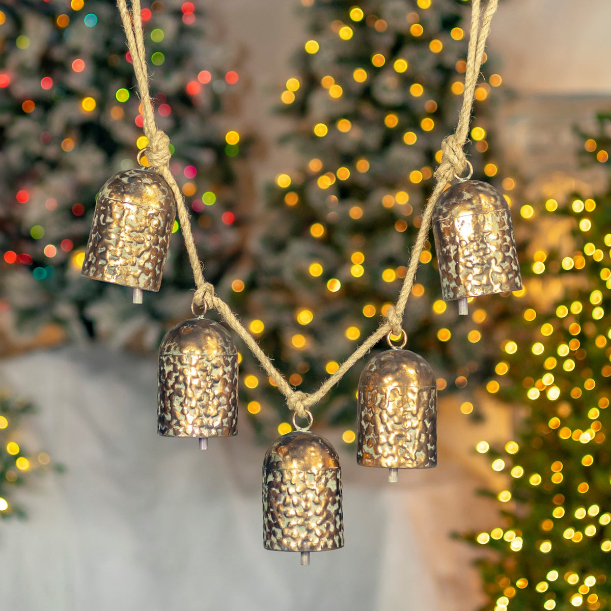 Five antique-gold textured bells hang from a rustic rope in front of a softly lit Christmas tree.