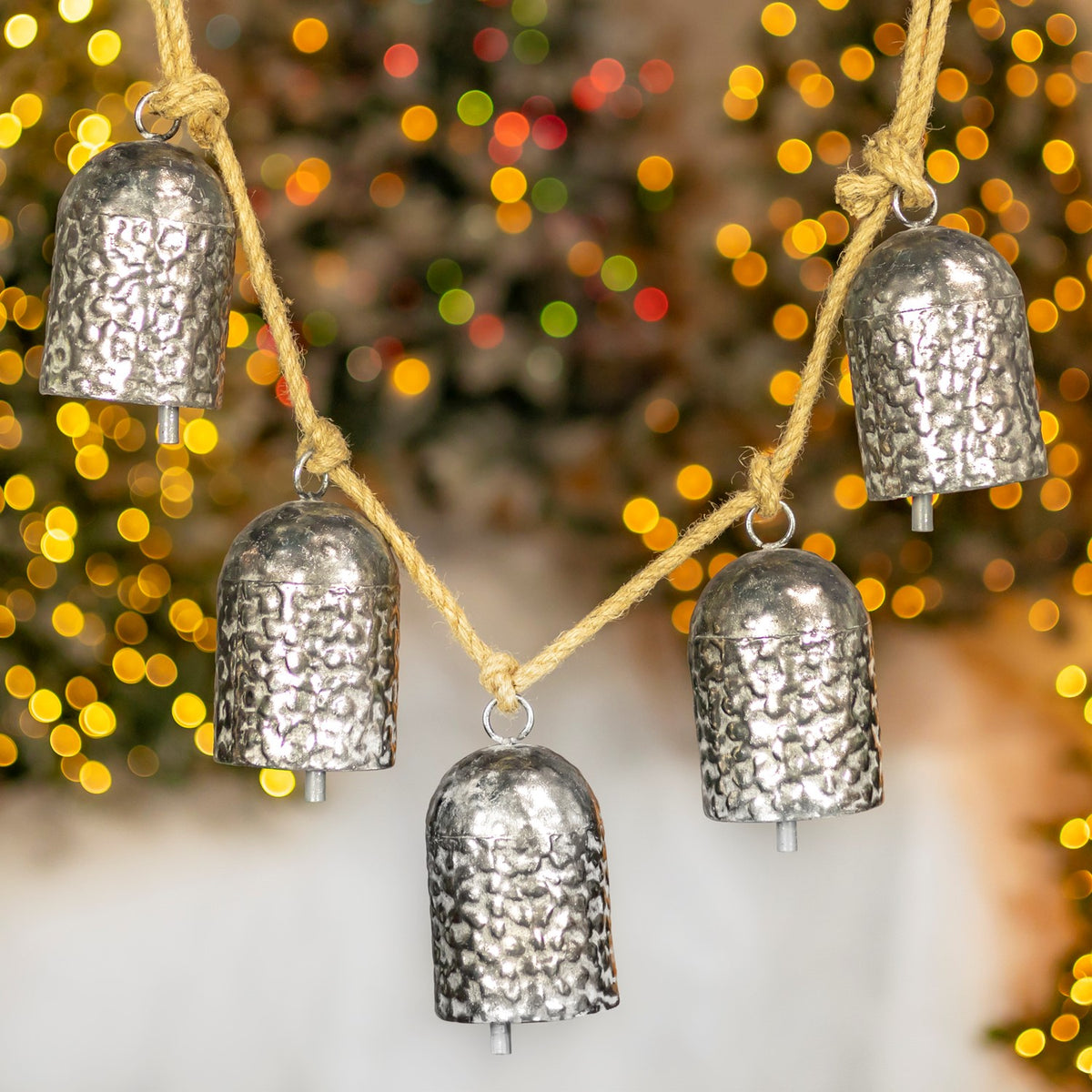 Five hammered silver bells hang from a coarse twine strand against warm, blurred lights.