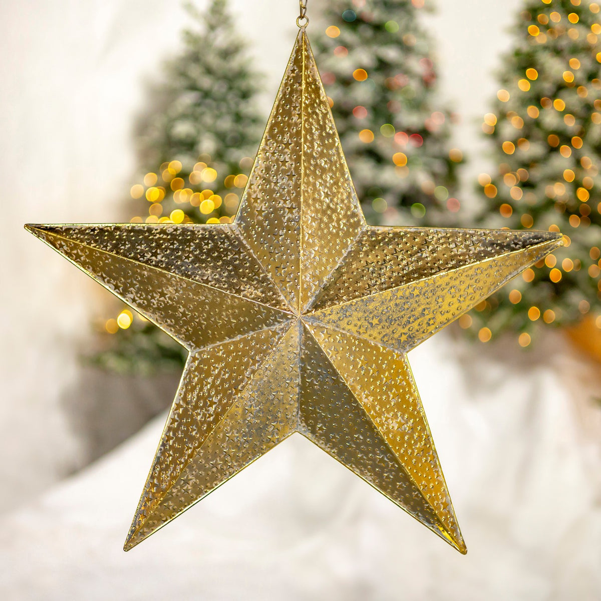 A textured gold five-point star ornament hangs in front of softly lit Christmas trees.