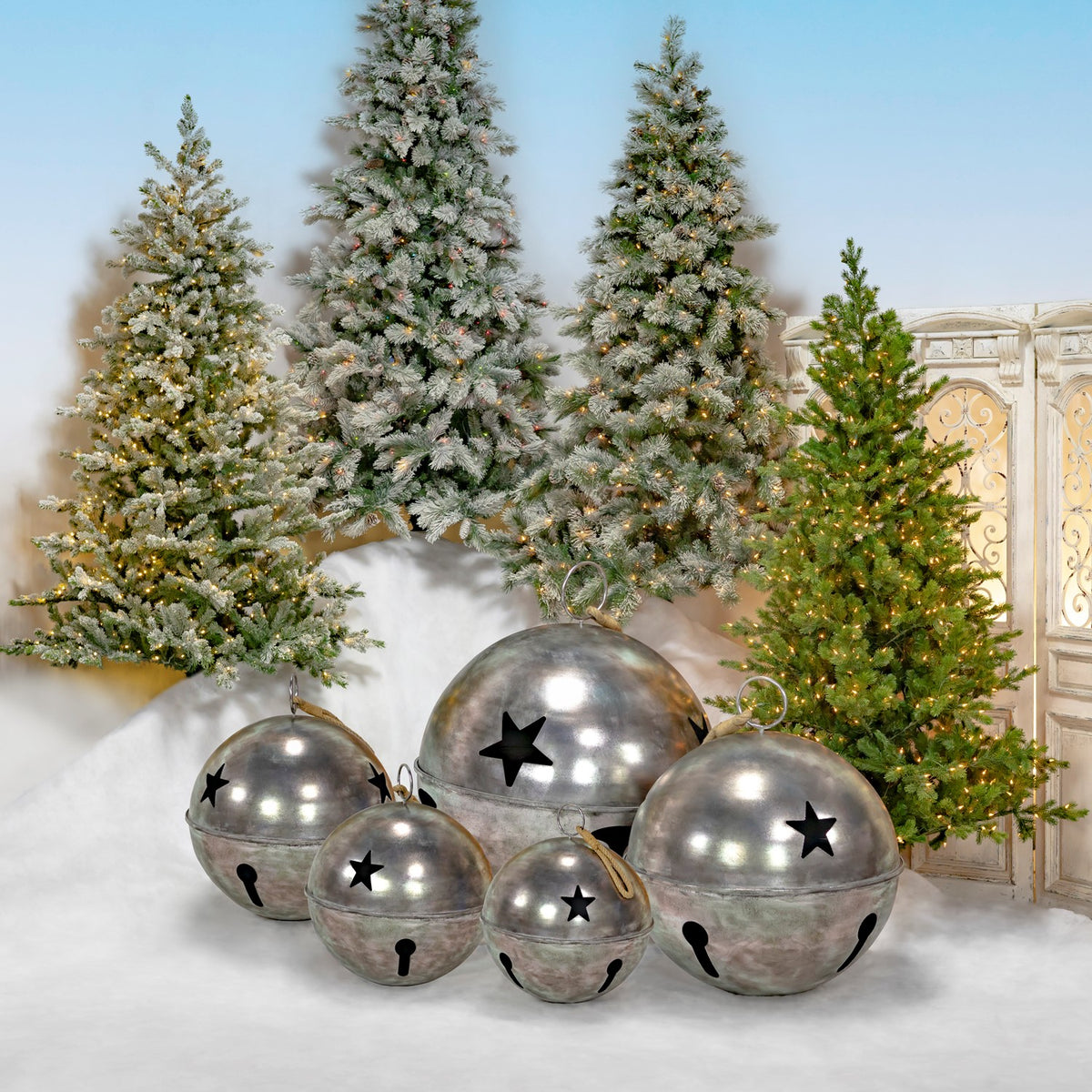 Five silver bells of different sizes with star cutouts arranged before decorated Christmas trees.