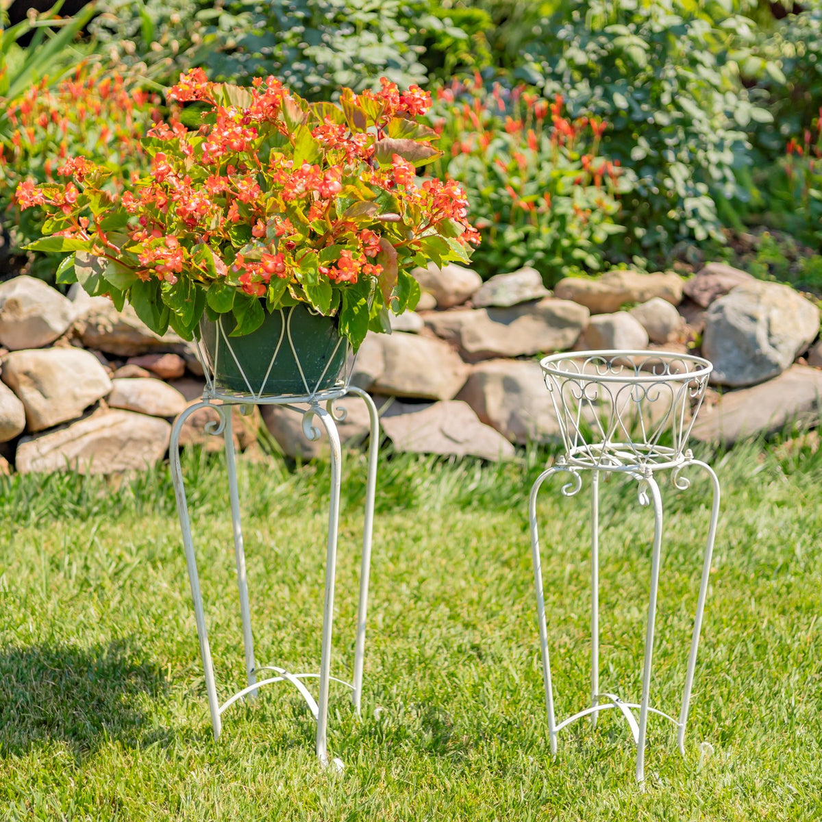 Two ornate white metal plant stands on grass, with flowers in one basket and the other empty.