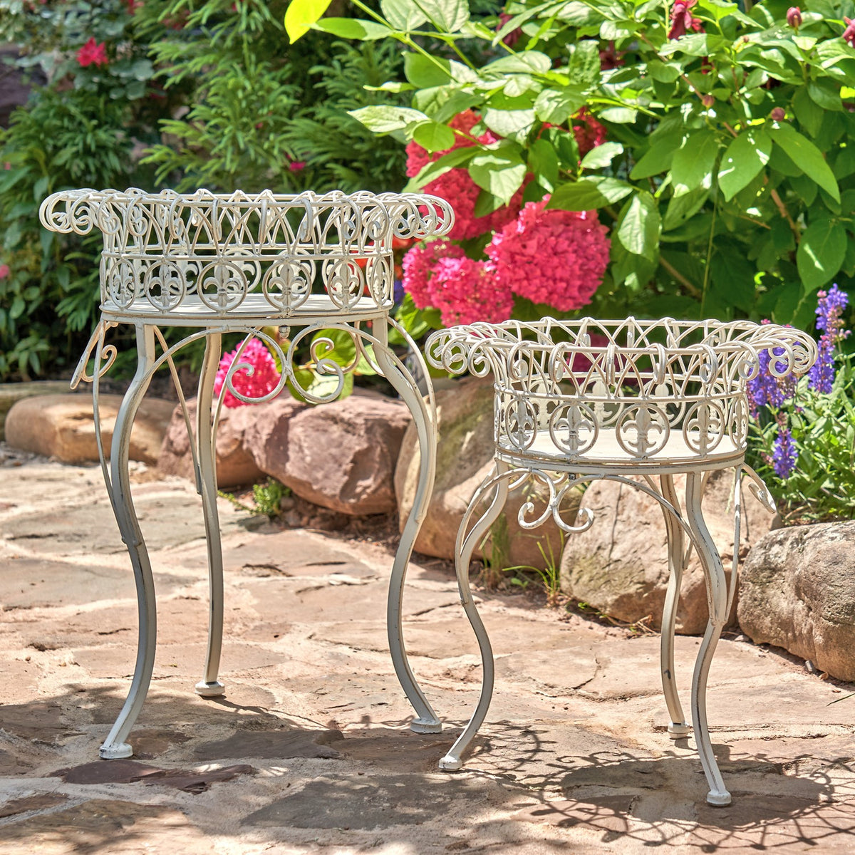 Two ornate light-colored metal plant stands with round basket tops on a stone patio among flowers.