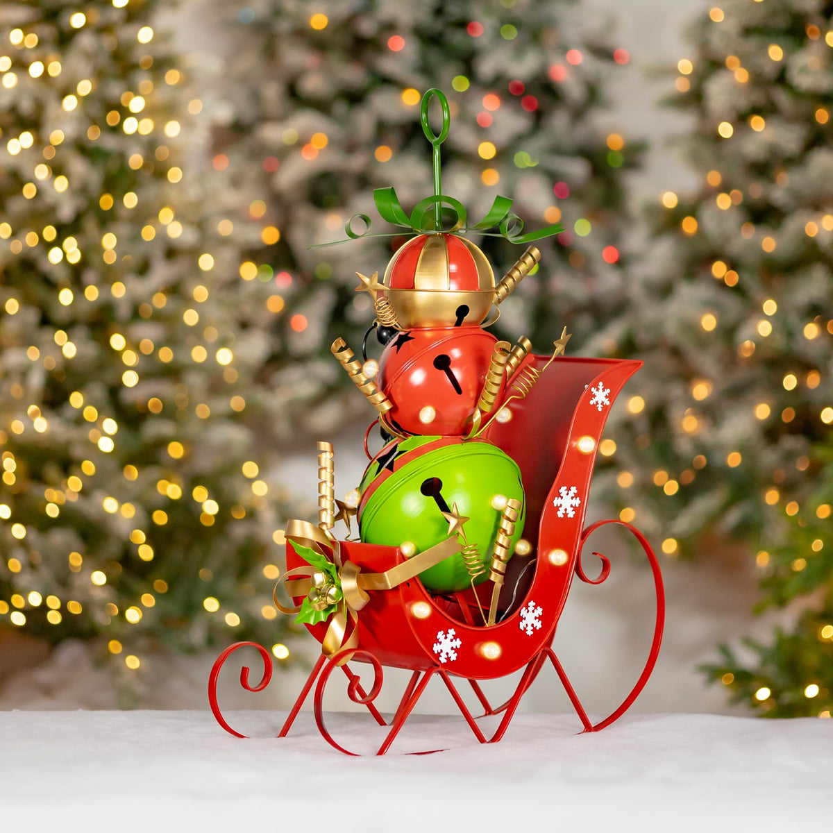 Red metal sleigh with a colorful stacked snowman figure, gold accents and bells on a snowy surface.