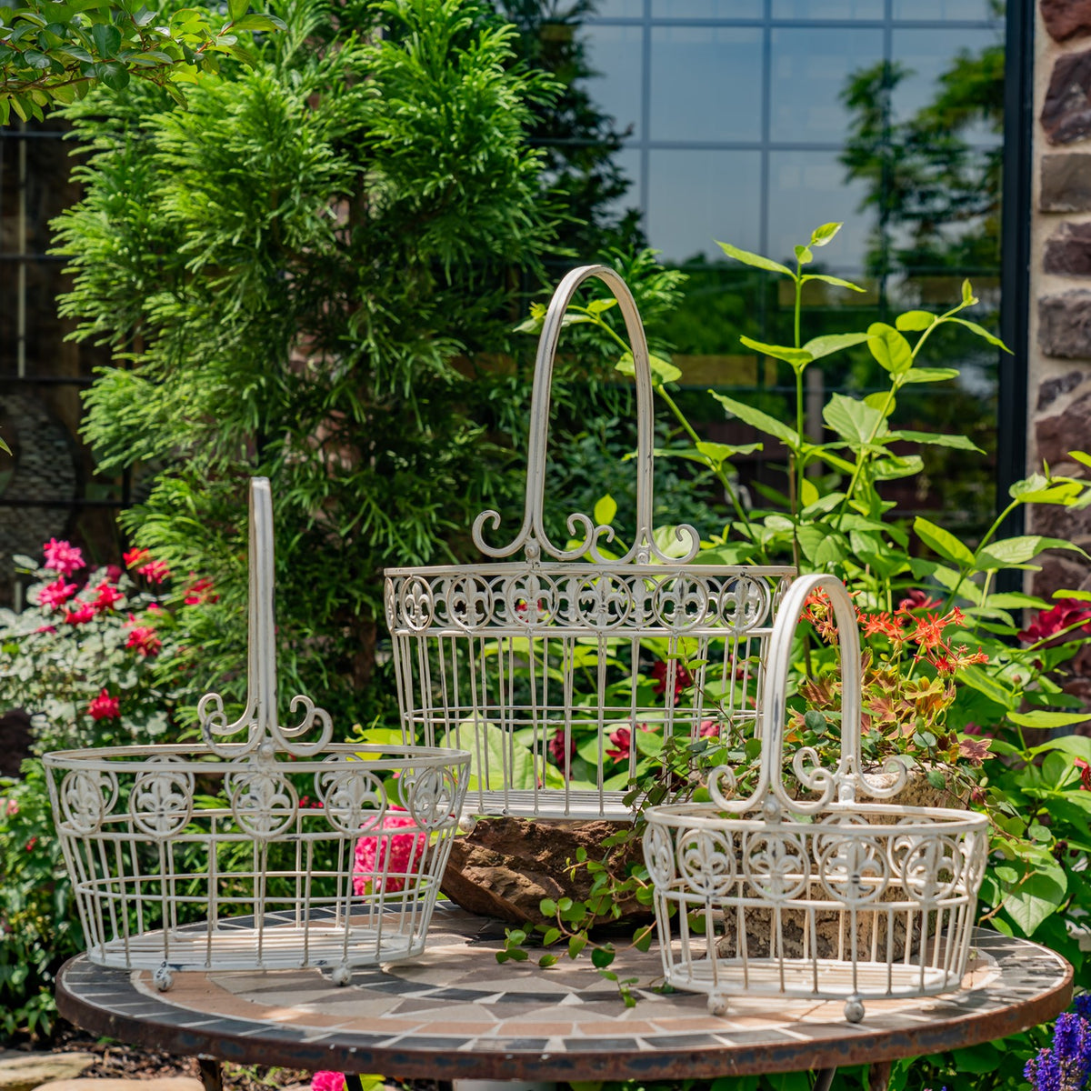 Three pale metal wire baskets with ornate rims and tall handles arranged on a garden table.