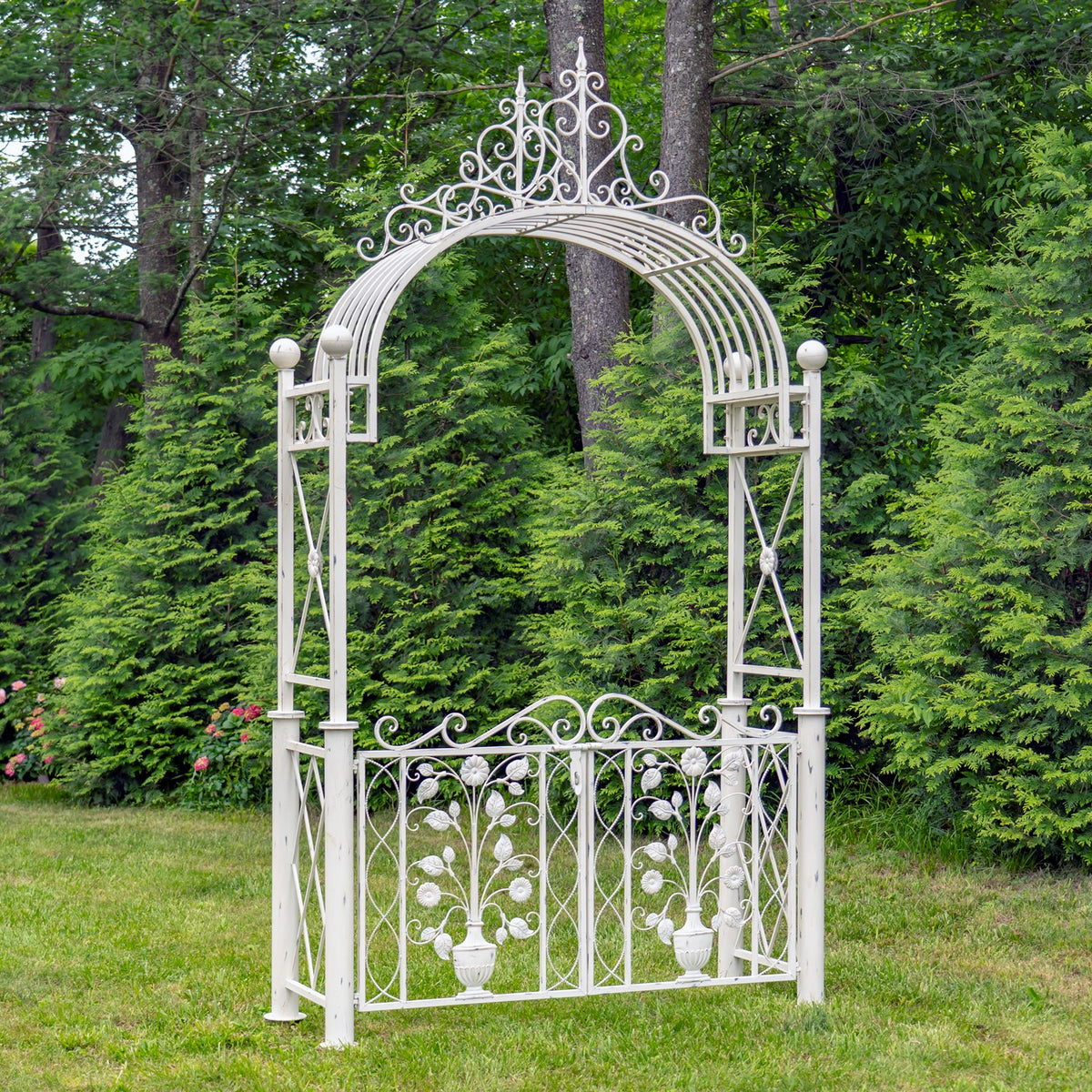 White ornamental garden arch and floral gate standing on a lawn among trees and shrubs.