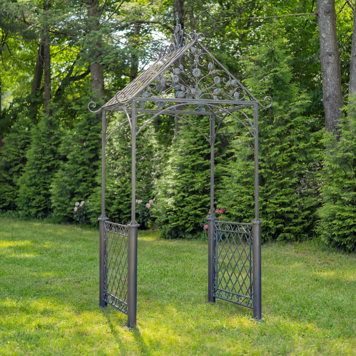 Ornate iron garden arch standing on a lawn, with trellis side panels and a peaked decorative roof.