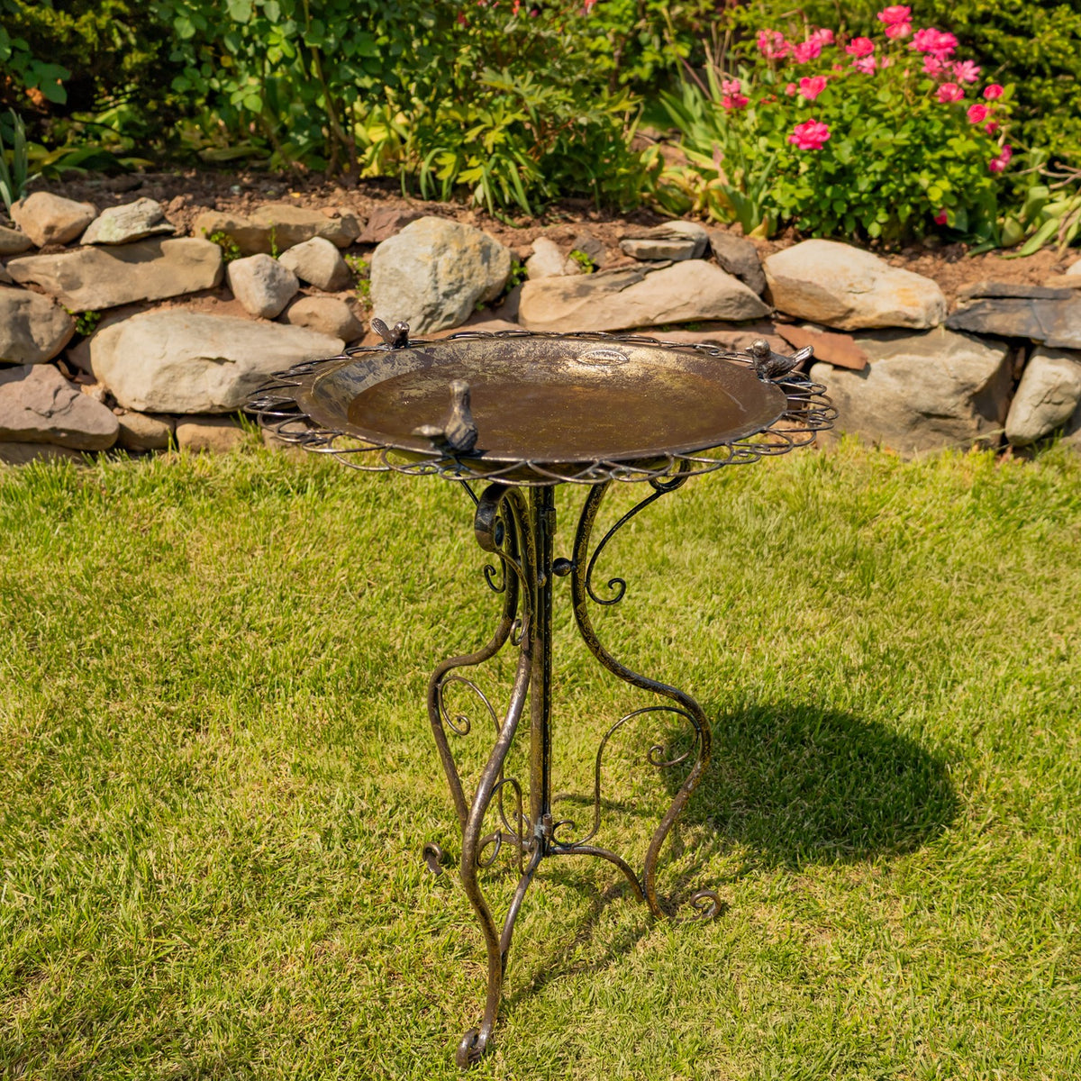 Round birdbath with decorative scrollwork legs sits on a lawn beside a low stone garden wall.