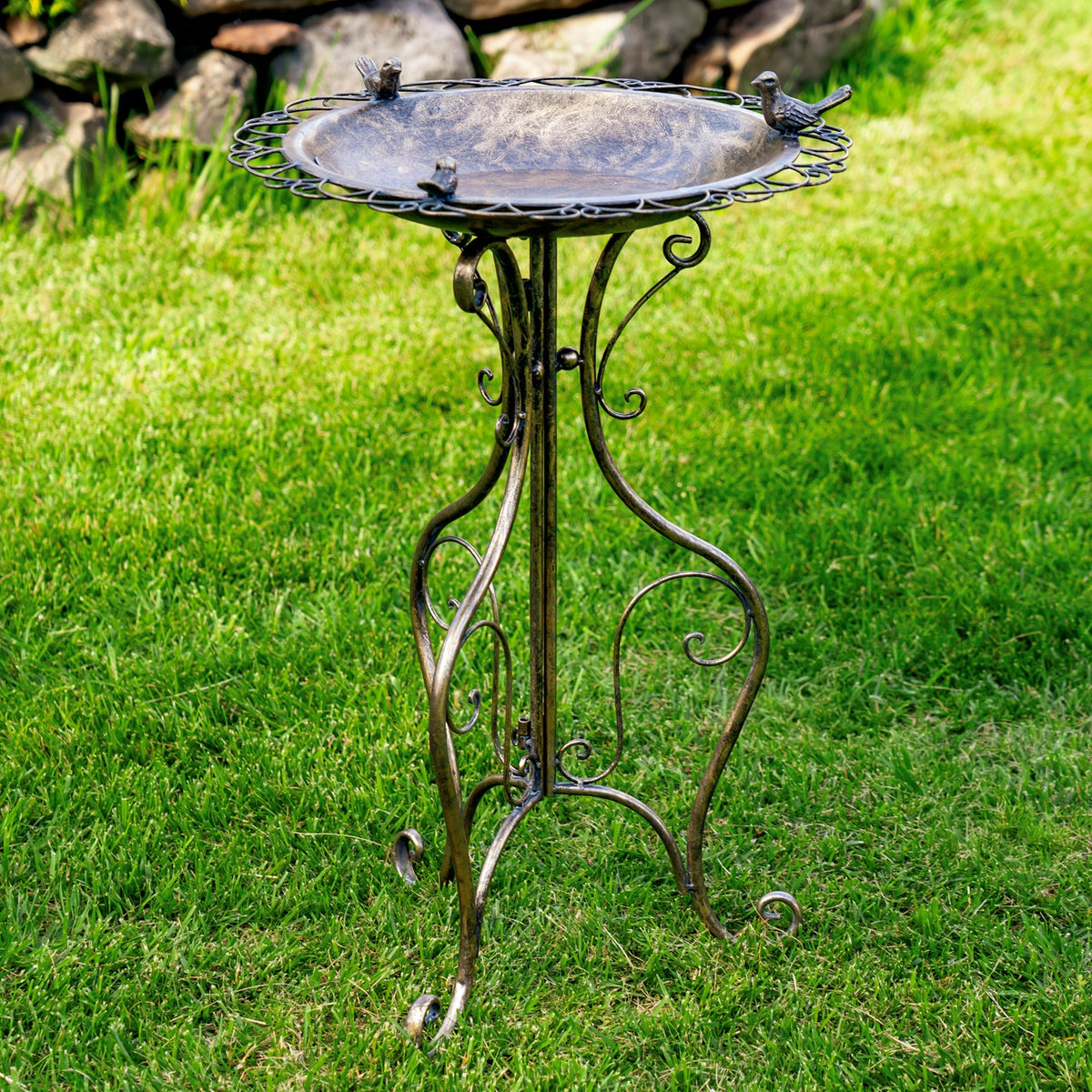 Ornate round metal birdbath on a curled iron stand in a grassy garden, with three small bird figures around the rim.