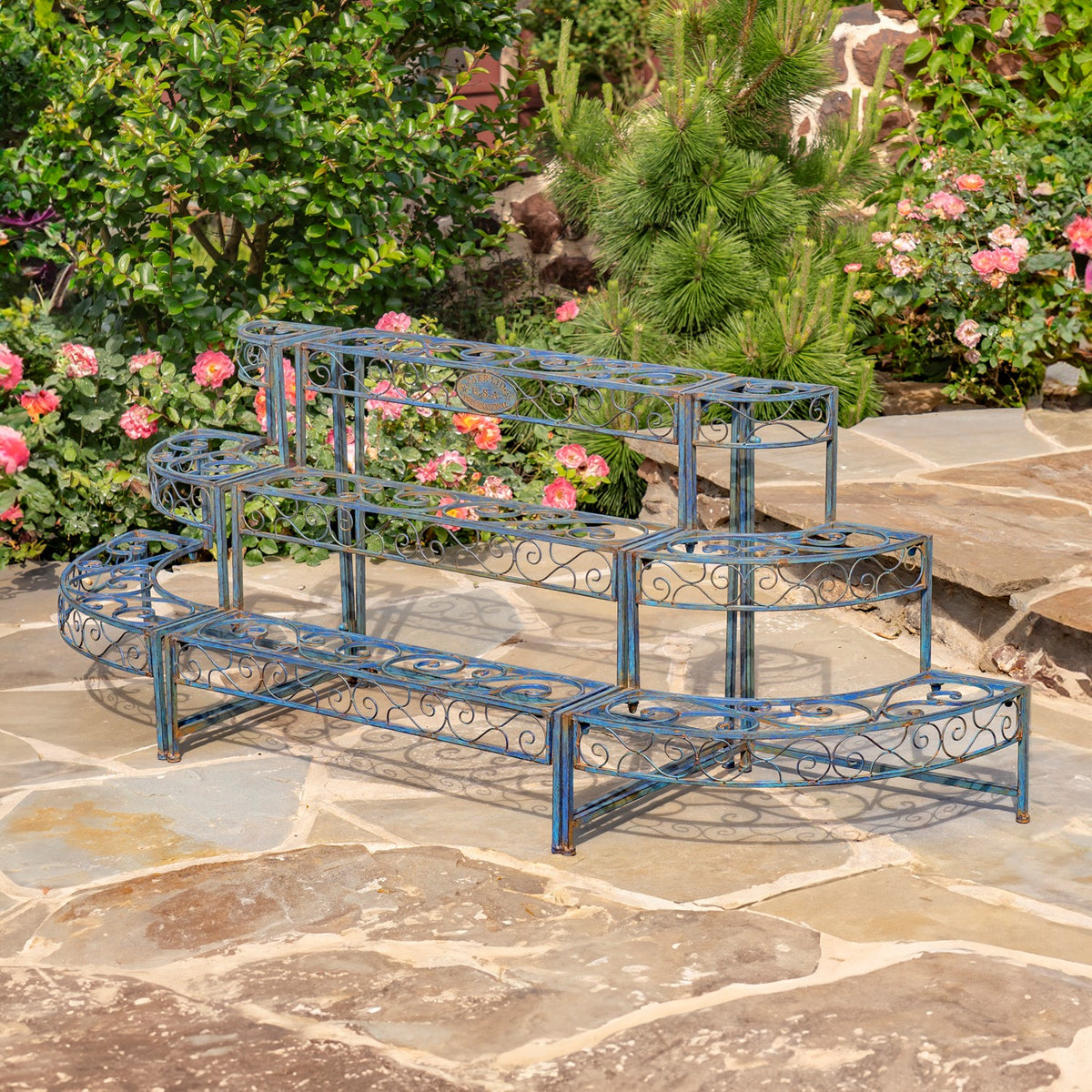 Three-tier blue metal plant stand with decorative scrollwork displayed on a stone patio among flowering plants.