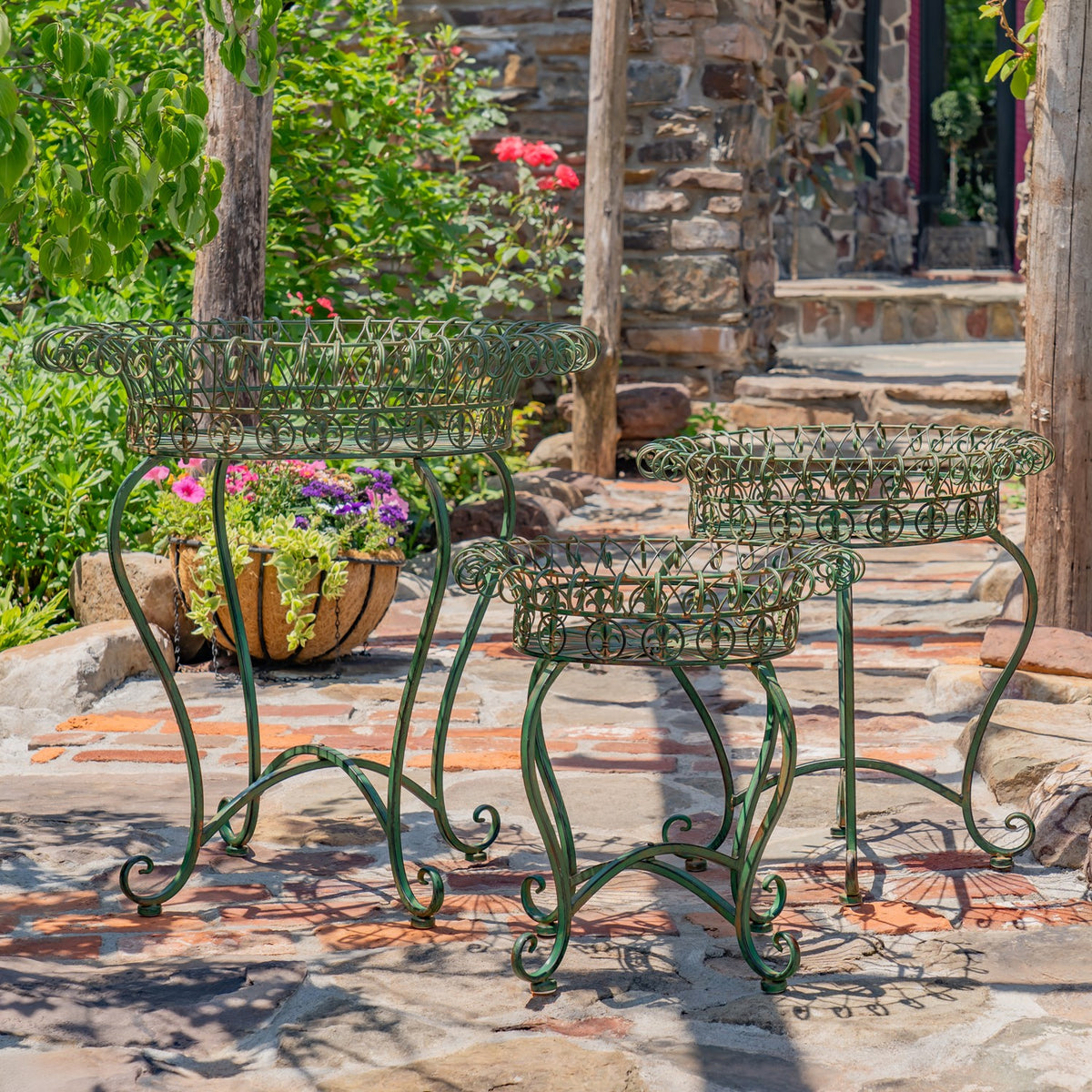 Three dark green wire basket stands arranged among plants along a stone walkway.
