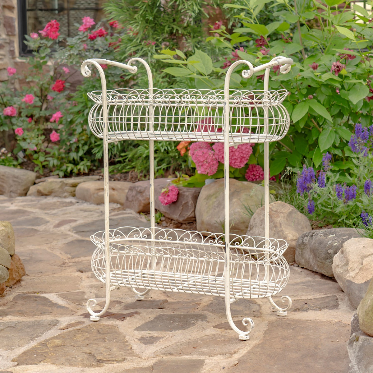 Antique-white metal plant stand with two oval wire baskets displayed among garden flowers.