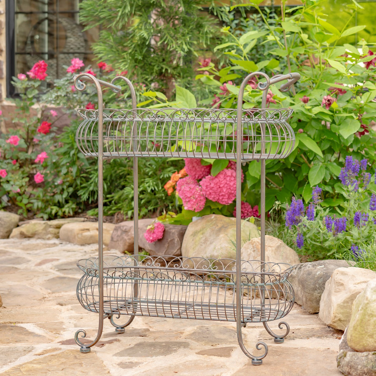 Two-tier oval wire plant stand with curled feet displayed among garden flowers and rocks.