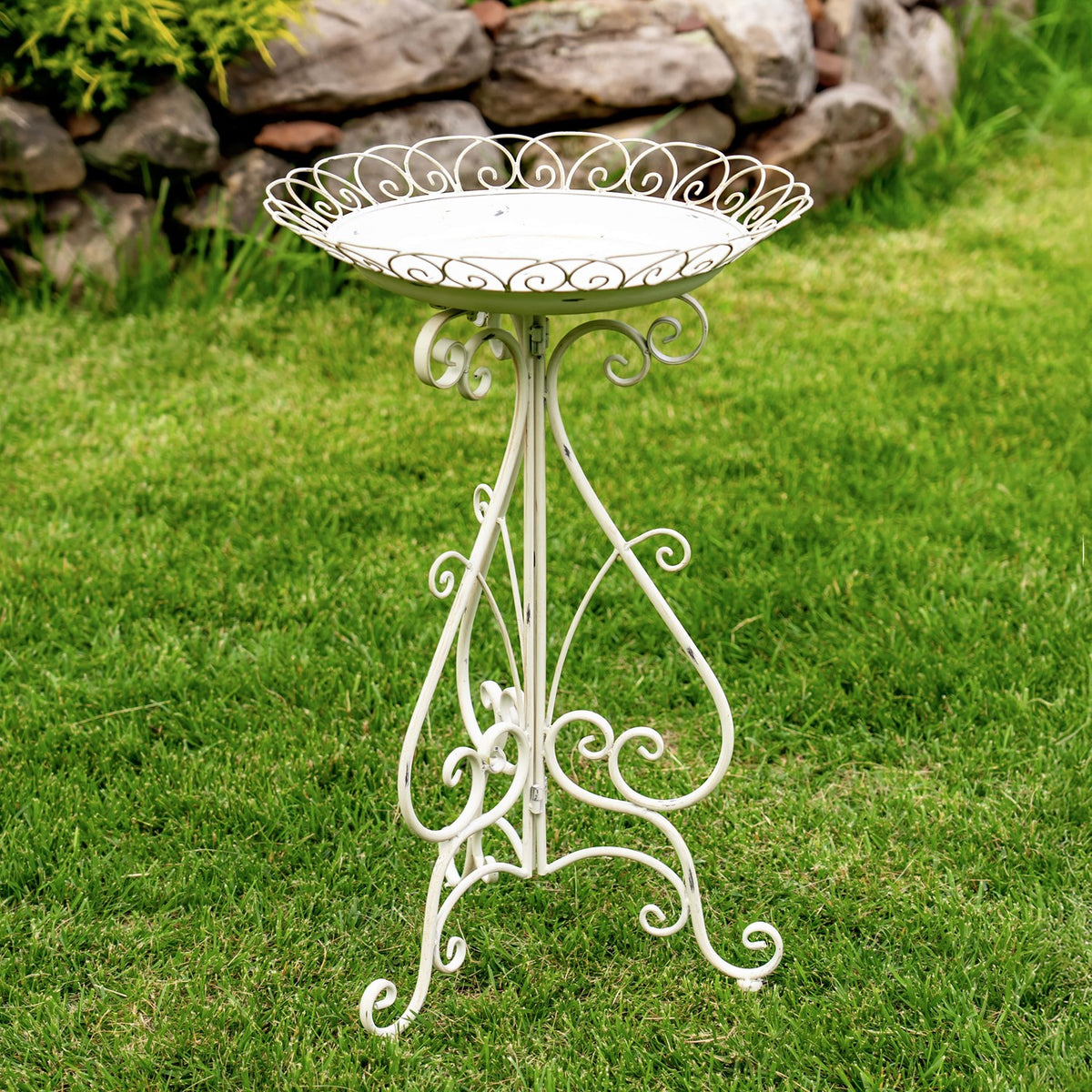 White scrollwork birdbath on a matching three-legged stand in a grassy garden.