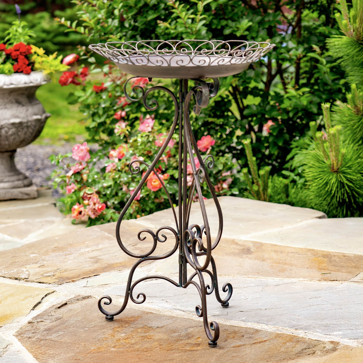 Ornate metal birdbath on a curled-leg stand, surrounded by flowers and greenery on a stone patio.