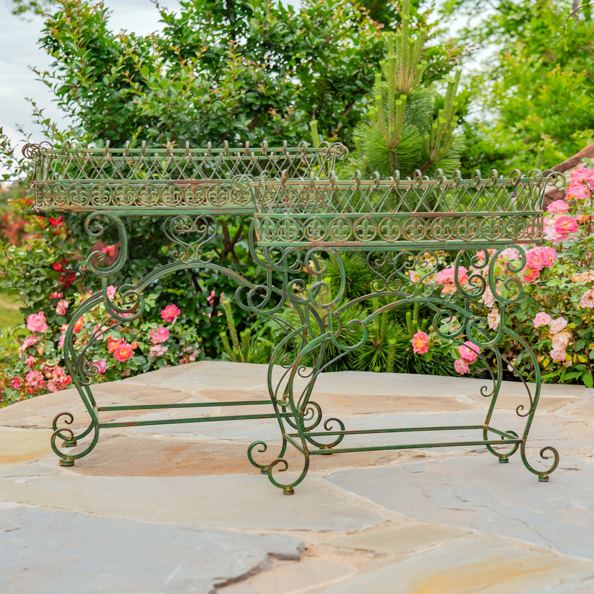Two dark green iron stands with embellished oval baskets, shown among flowering plants.