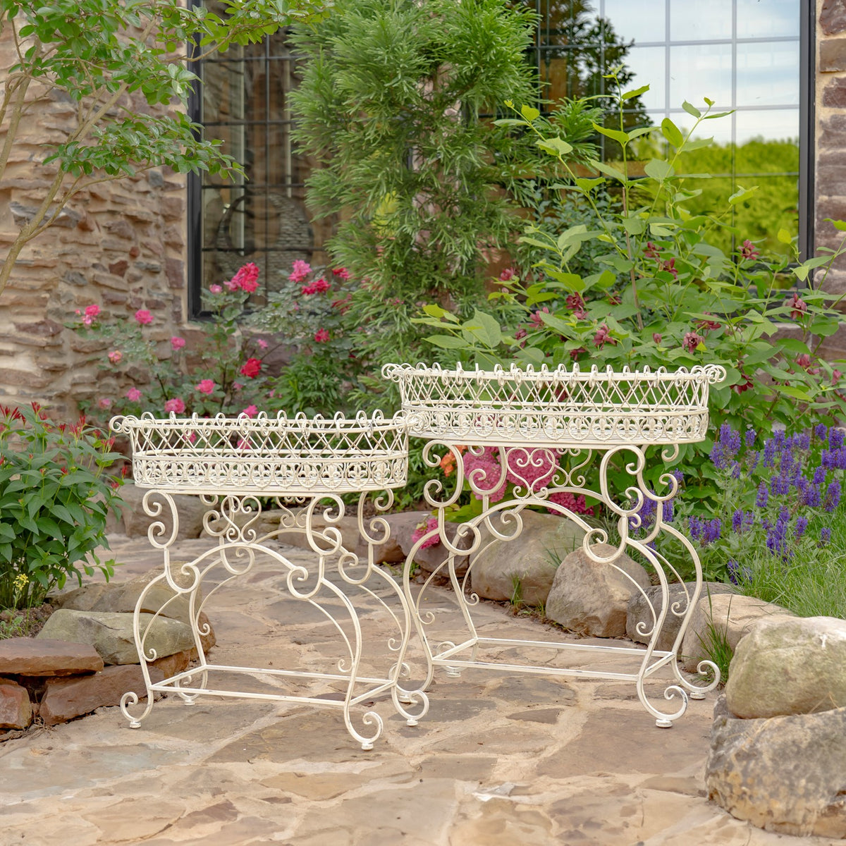 Two white ornate iron plant stands with oval baskets arranged on a stone patio among flowers.