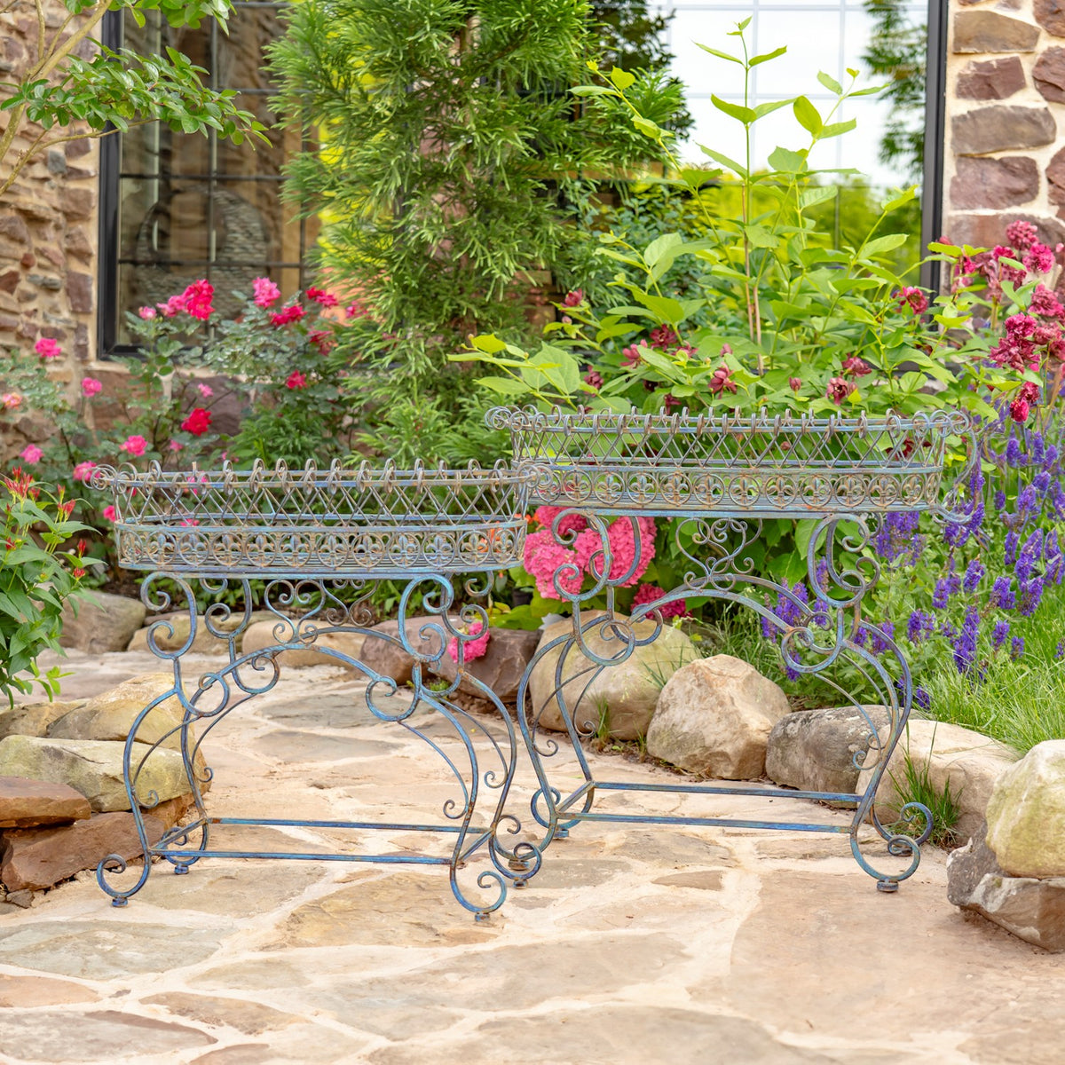 Pair of blue scrollwork iron plant stands with oval baskets displayed in a garden setting.