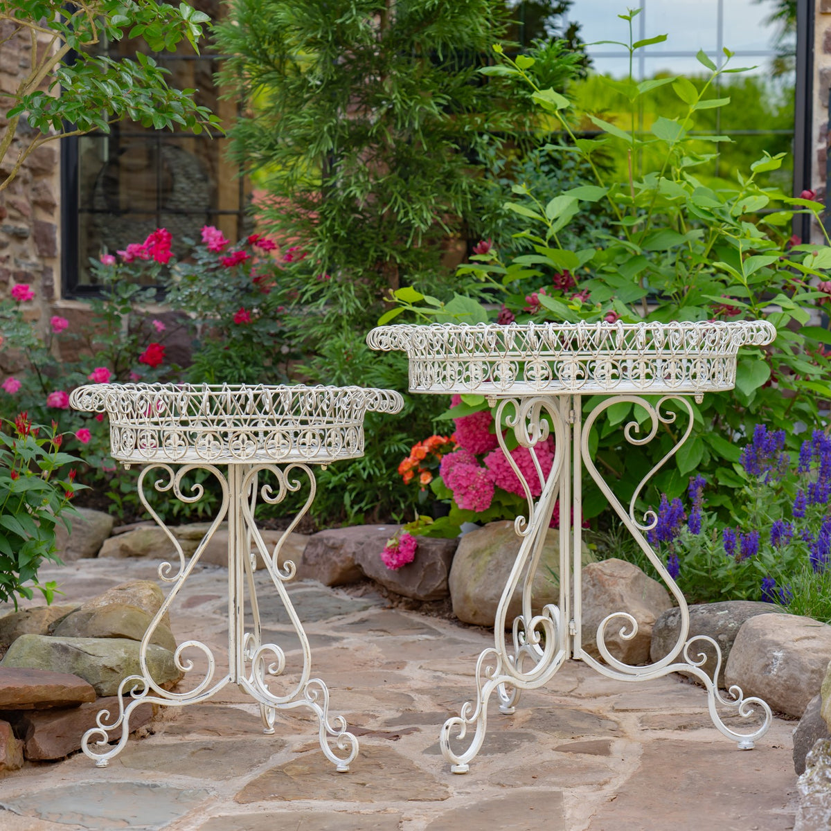 Two white round wire-basket plant stands on a stone garden path among flowering plants.