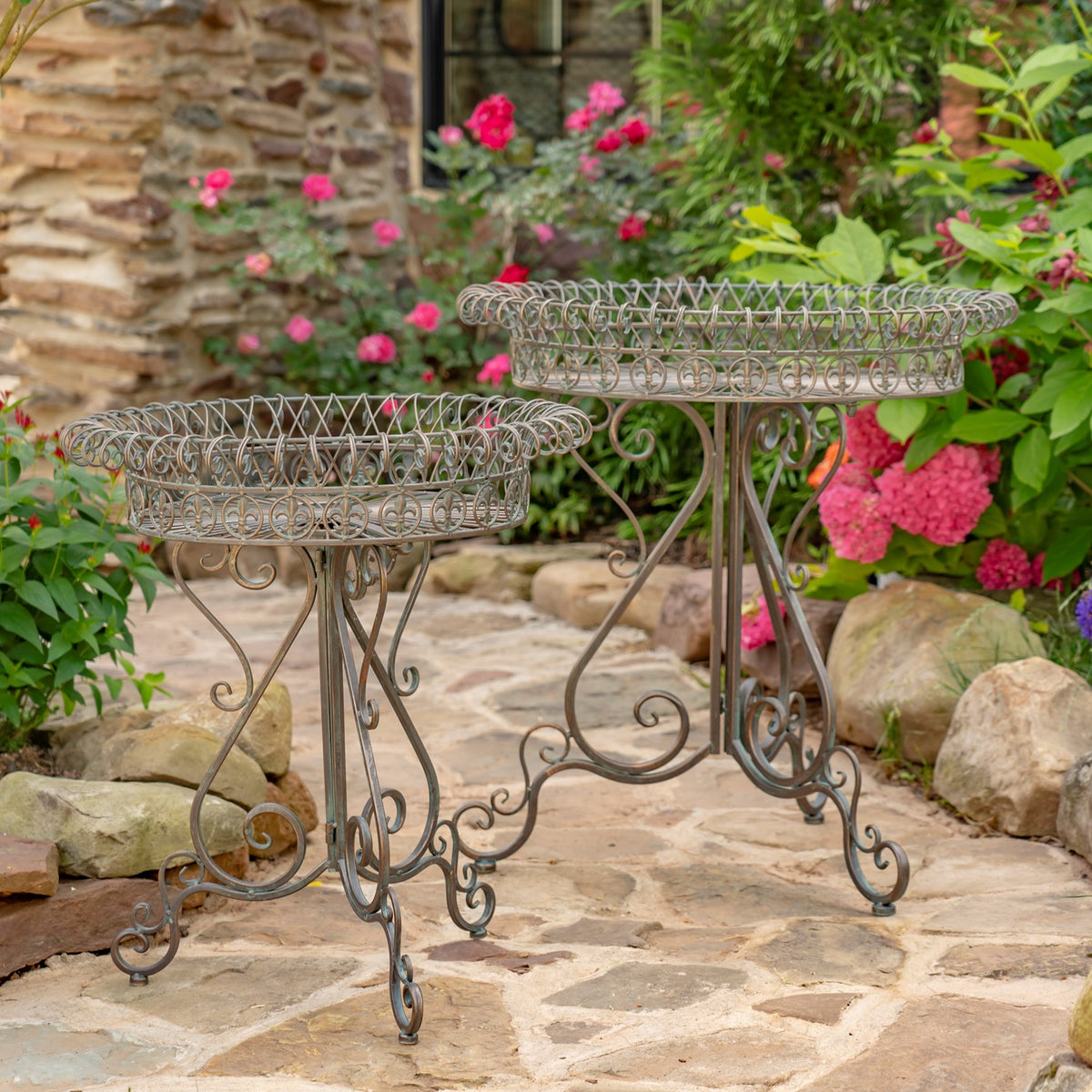 Two dark metal plant stands with round decorative wire baskets in a garden setting.