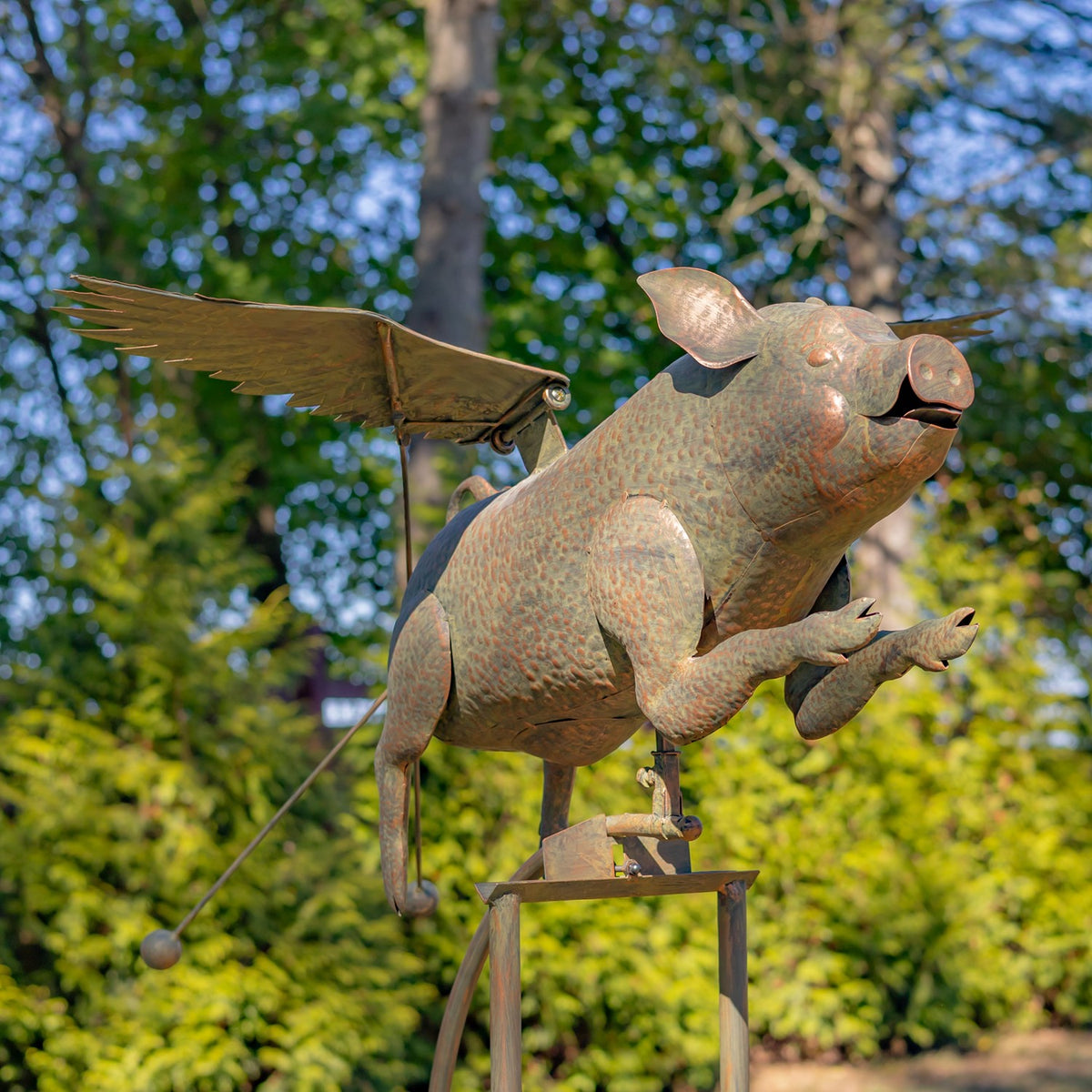Rustic metal pig with outstretched wings perched on a tall iron stake in a garden.