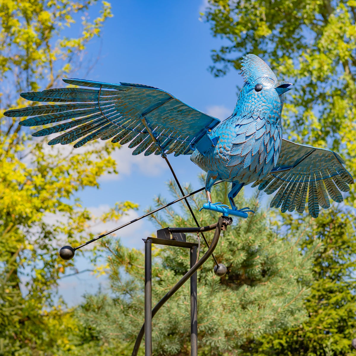 Blue metal jay with outstretched wings perched on a tall rocking stake among trees.