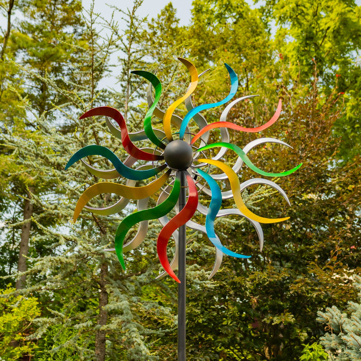 Colorful metal wind spinner with curled blades on a dark stake, shown among trees.