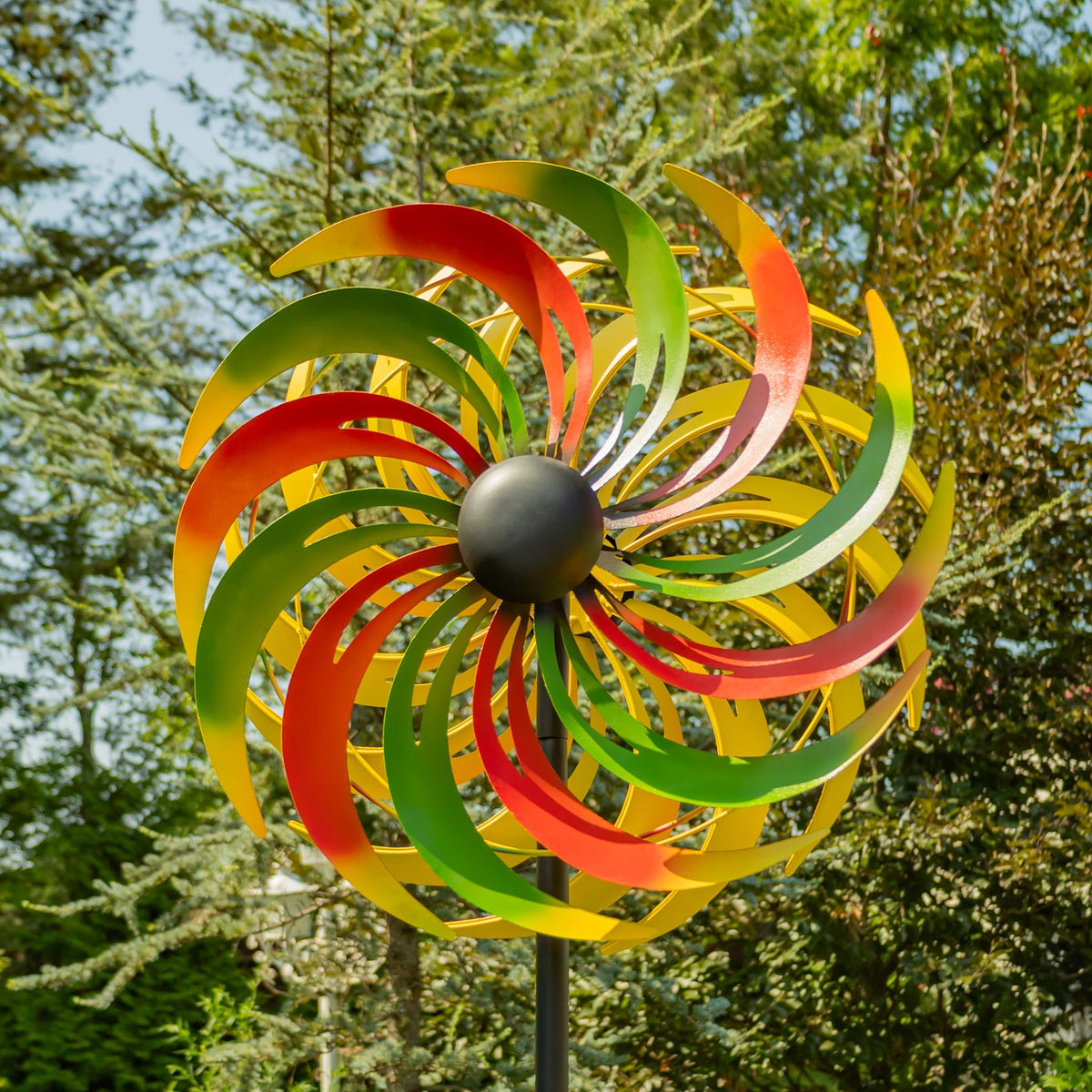 Red, green, and yellow curved metal blades spiral around a dark center against leafy trees.