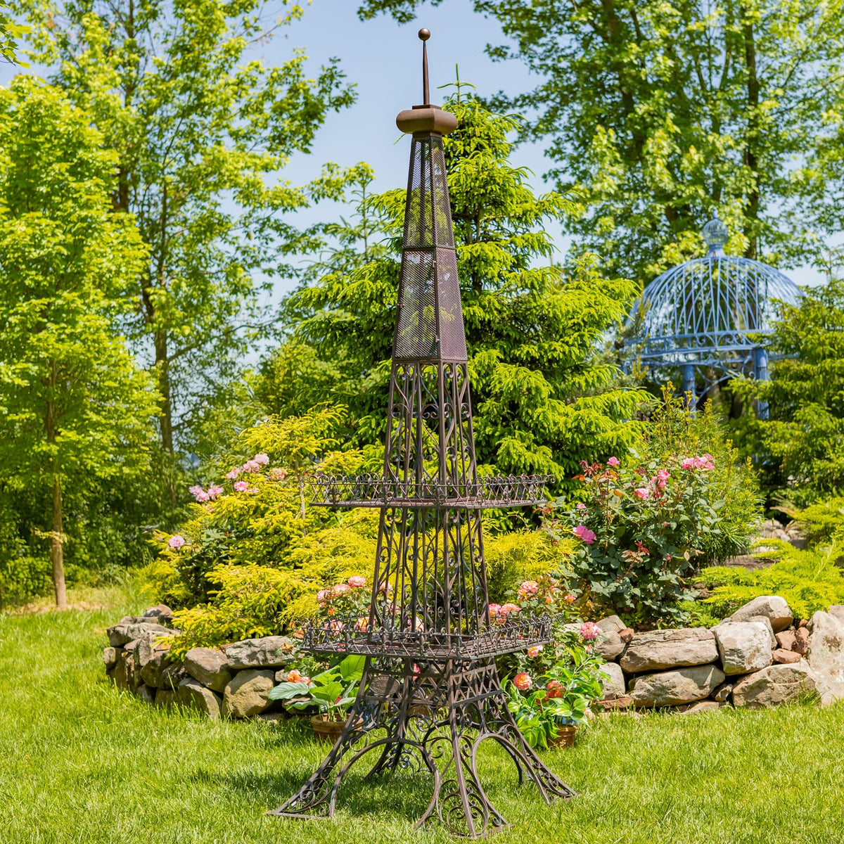 Dark-finished Eiffel Tower-shaped plant stand with two tiers displayed beside flowering plants outdoors.