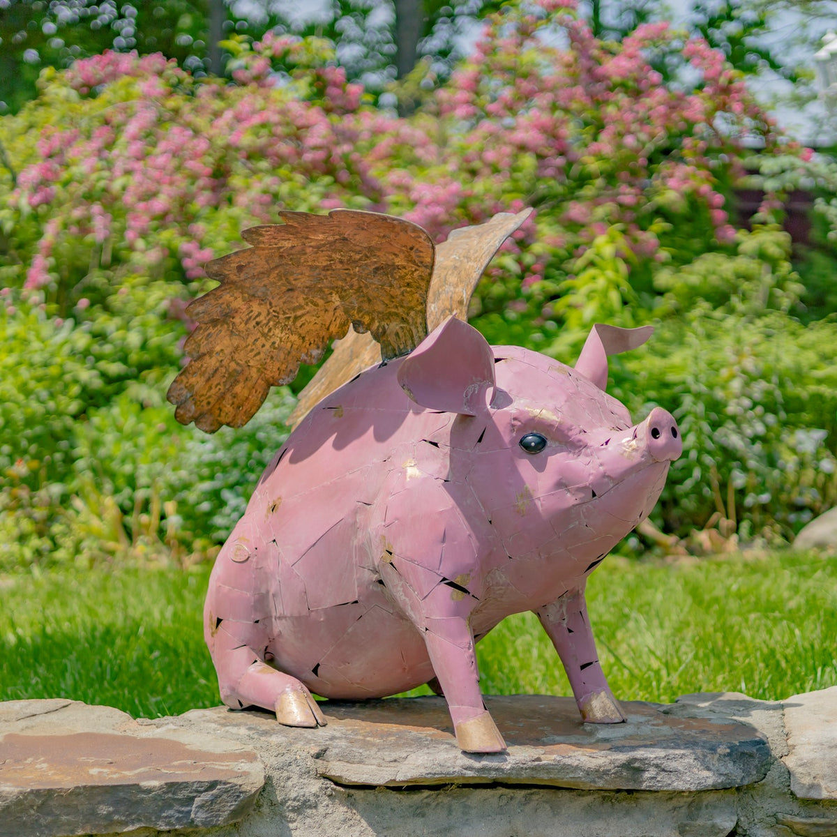 Pink metal pig statue with large weathered gold-toned wings standing on a stone ledge in a garden.