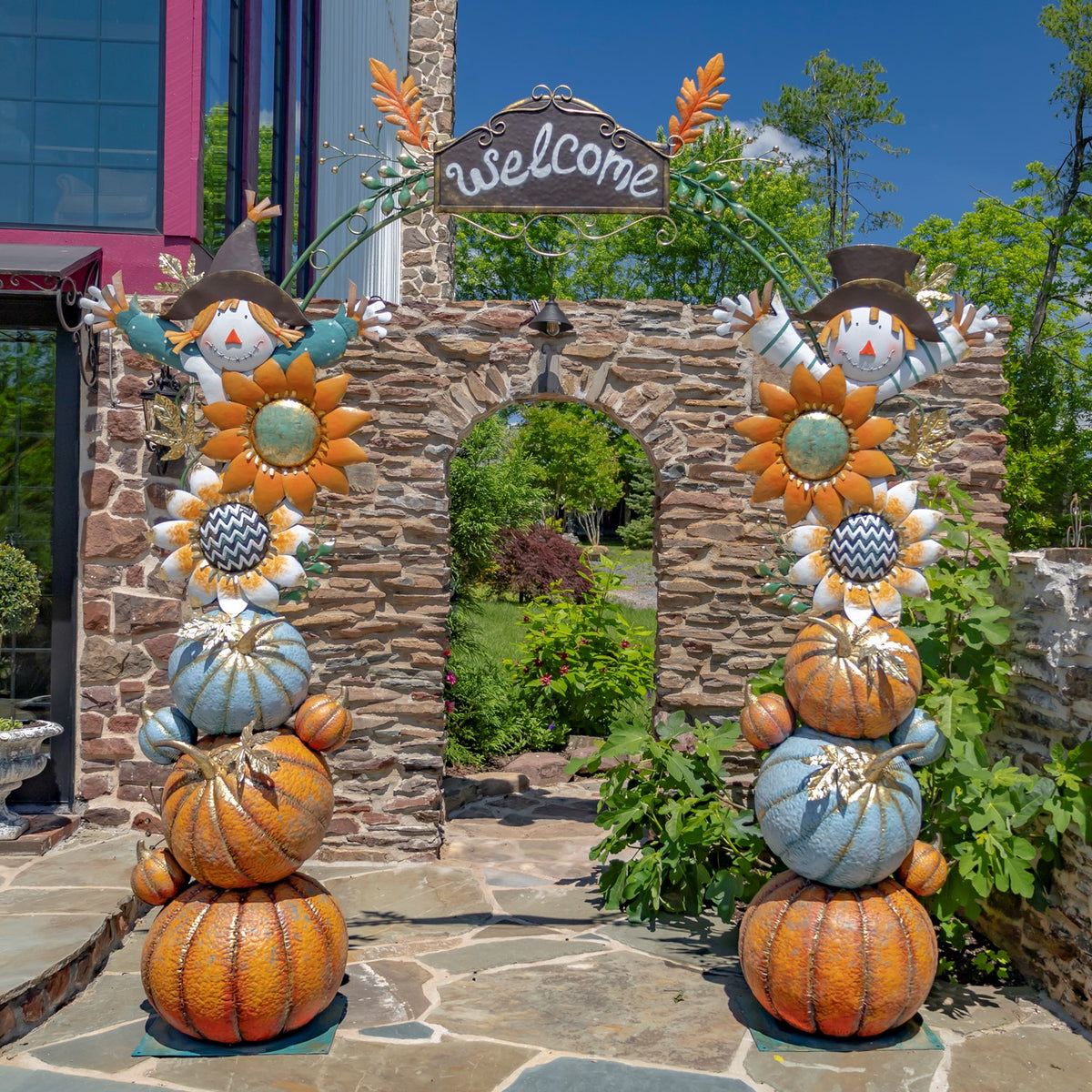 A harvest arch with matching pumpkin pillars, sunflower accents, and scarecrow figures frames a stone garden entrance.