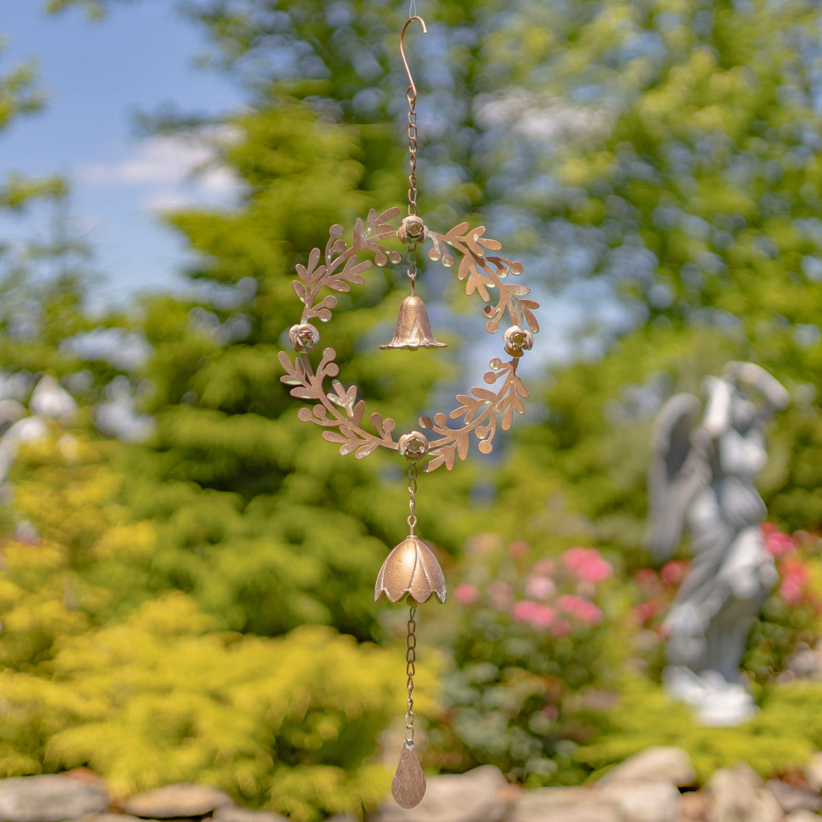 Copper-toned leafy wreath with dangling bells and a teardrop charm hangs against a garden backdrop.