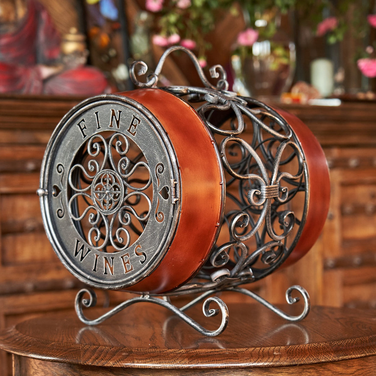 Oval wine rack with ornate metal scrollwork and reddish-brown bands, resting on curled feet on a wooden table.