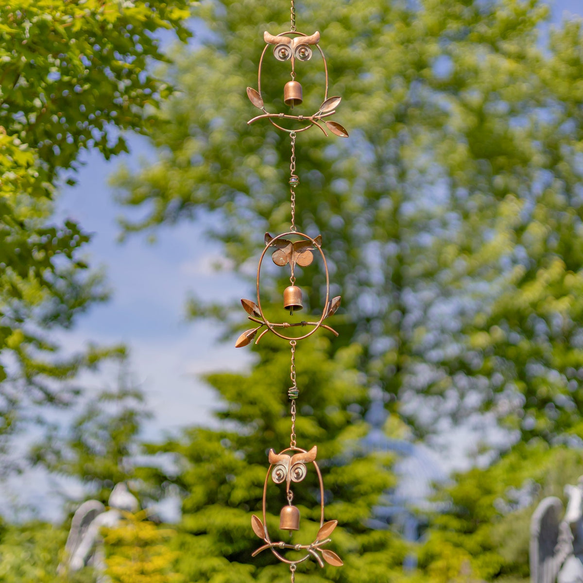 Copper-toned rain chain with repeating owl and leaf shapes hanging among green trees.