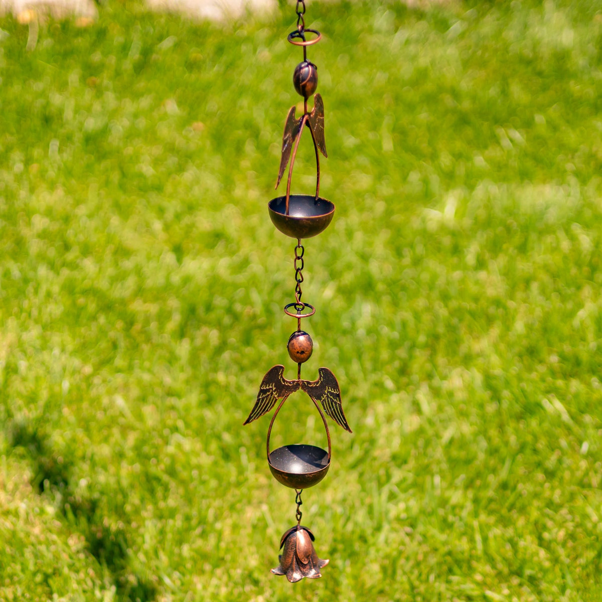 Antique bronze rain chain with angel figures and shallow bowls hanging against a green lawn.