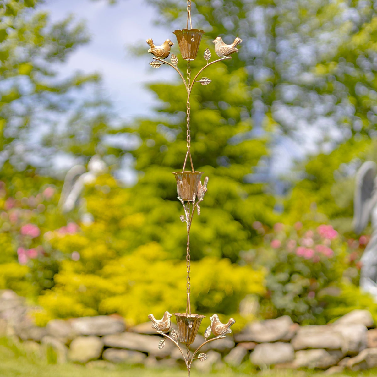 Antique copper rain chain with bird-and-flower ornaments hanging against a garden and stone wall.