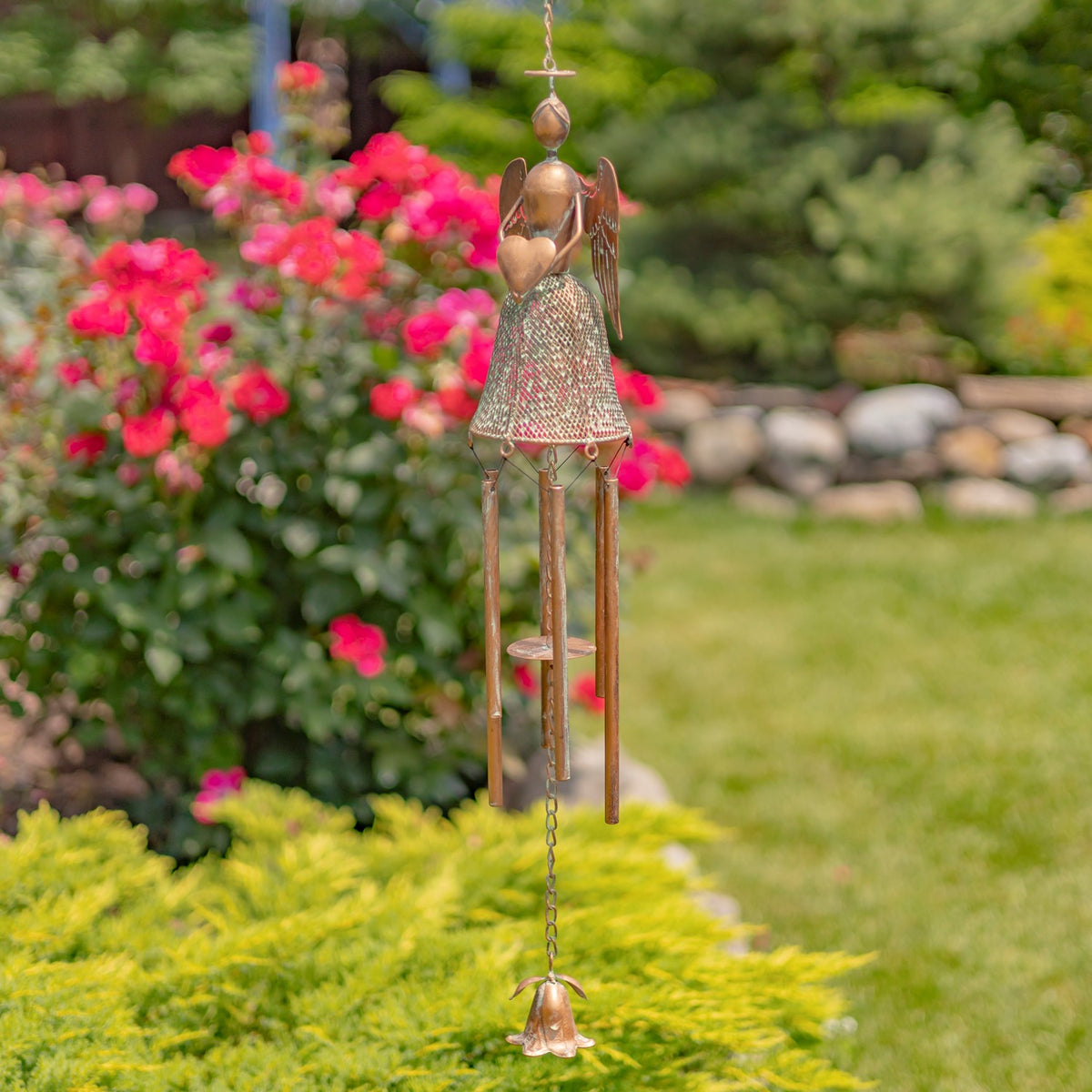 An angel holding a heart tops a hanging wind chime, shown among garden flowers and greenery.