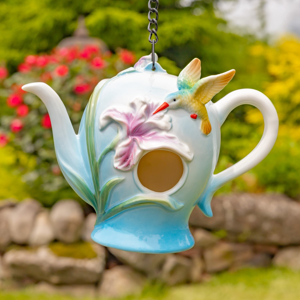 Blue teapot-shaped birdhouse with a hummingbird and flower hanging from a chain outdoors
