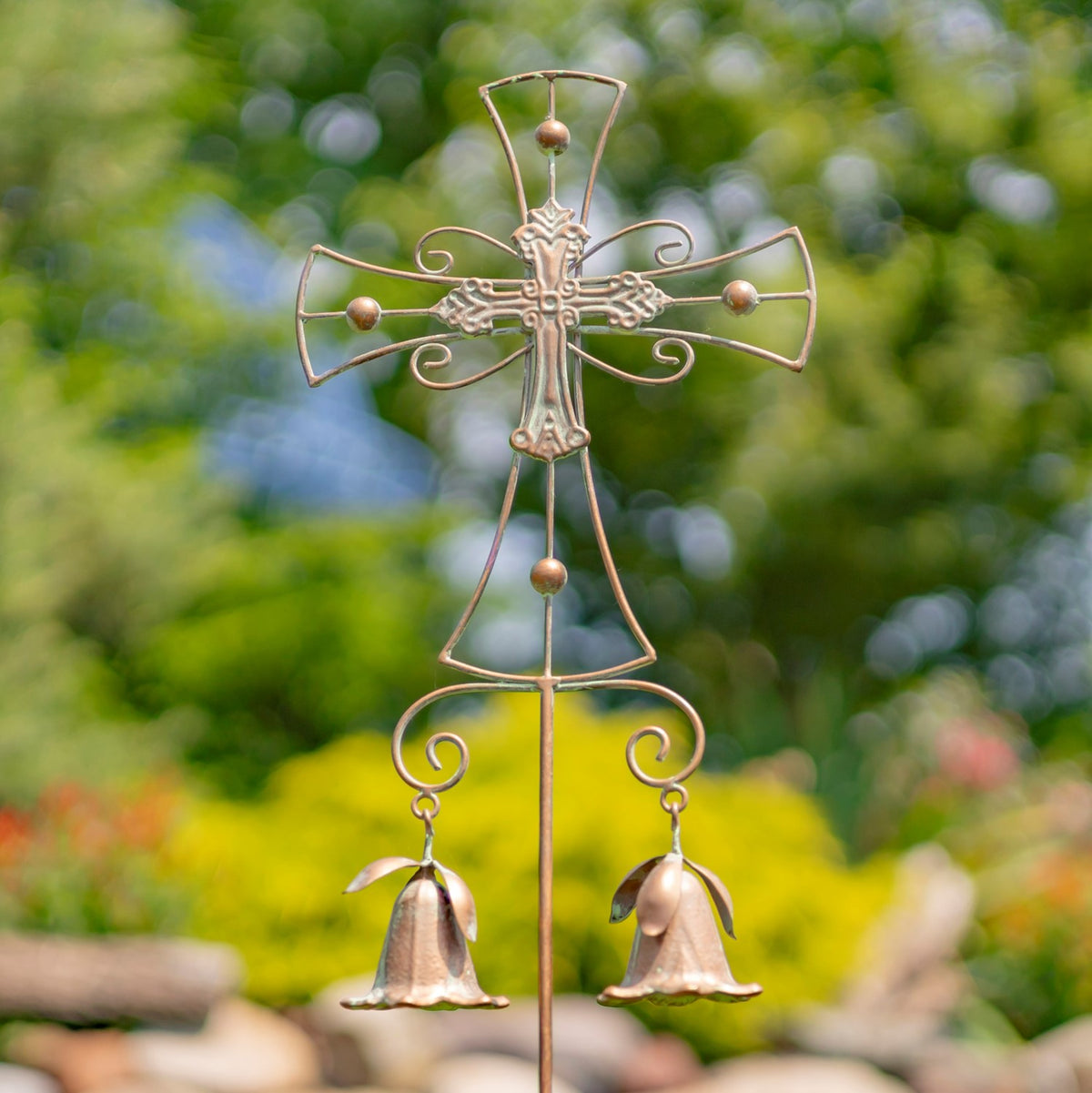 Close view of an ornate metal cross garden stake with two hanging bells against blurred greenery.