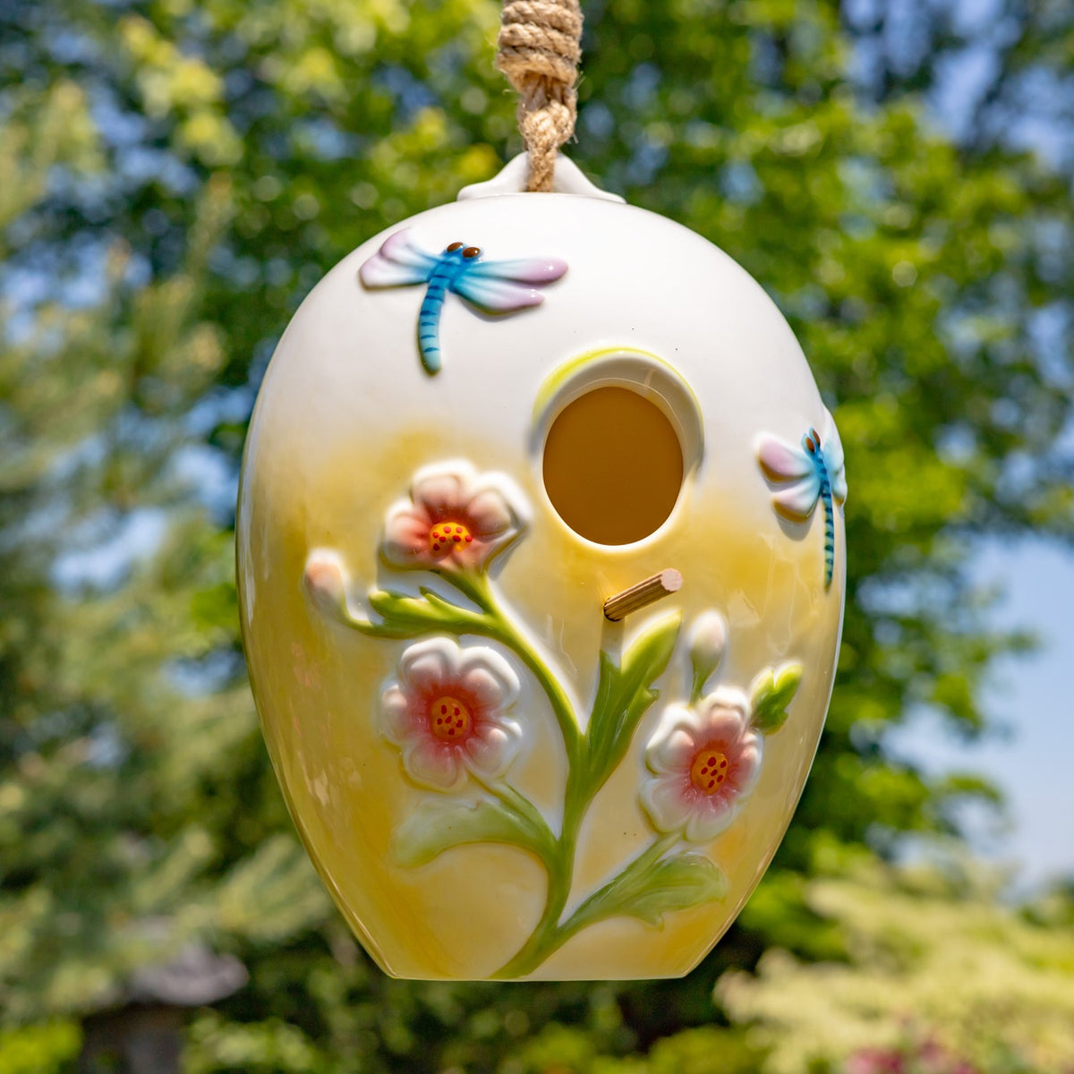 Egg-shaped birdhouse hanging outdoors, decorated with raised flowers and blue dragonflies around its entrance.