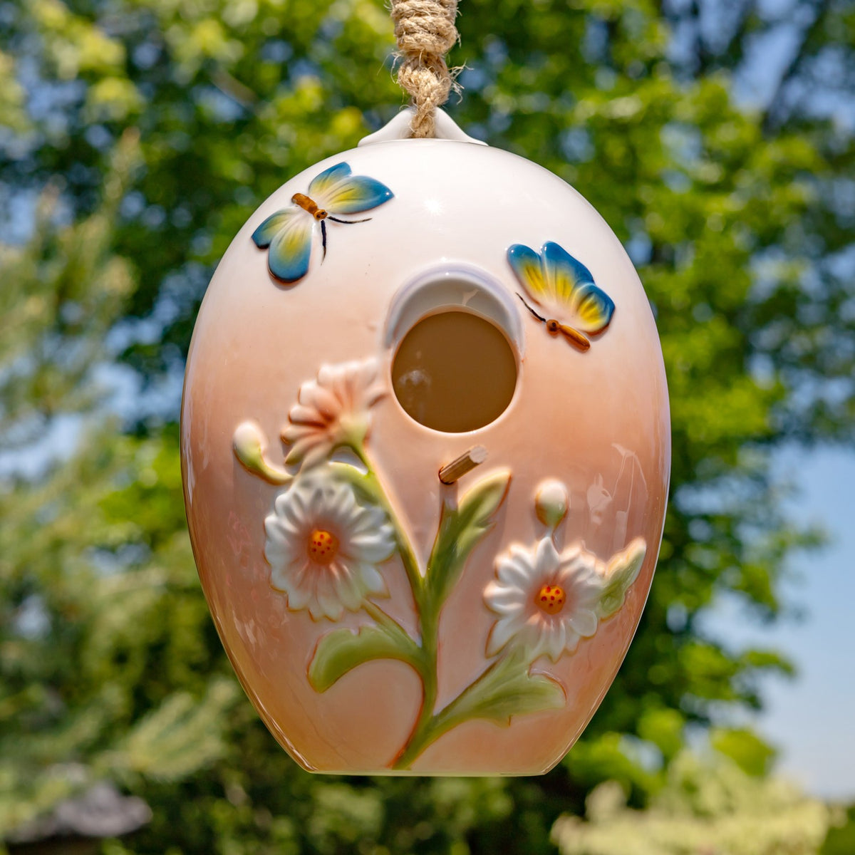 Egg-shaped hanging birdhouse with raised flowers and colorful butterflies against leafy trees.