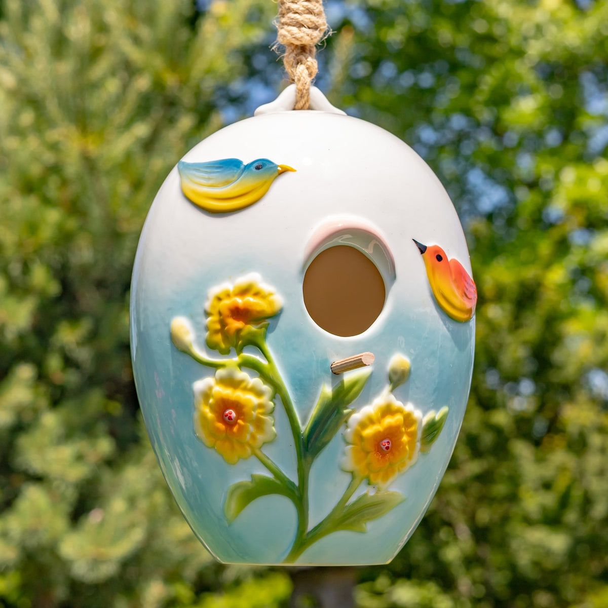 Egg-shaped birdhouse hanging outdoors, decorated with raised birds and yellow flowers around its round entrance.