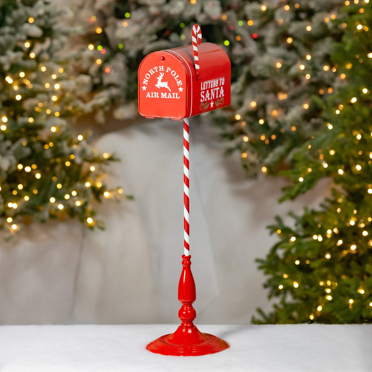 Red mailbox on a red-and-white striped stand, displayed in front of lit Christmas trees.