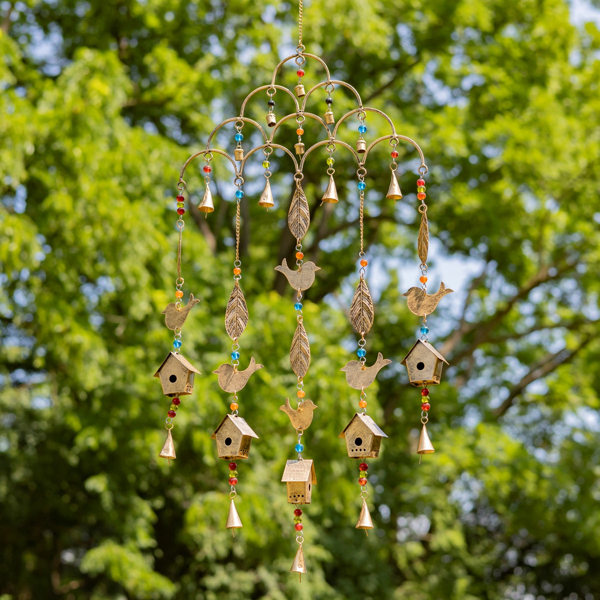 Gold-tone wind chime with dangling bird, leaf, bell and birdhouse ornaments against leafy trees.