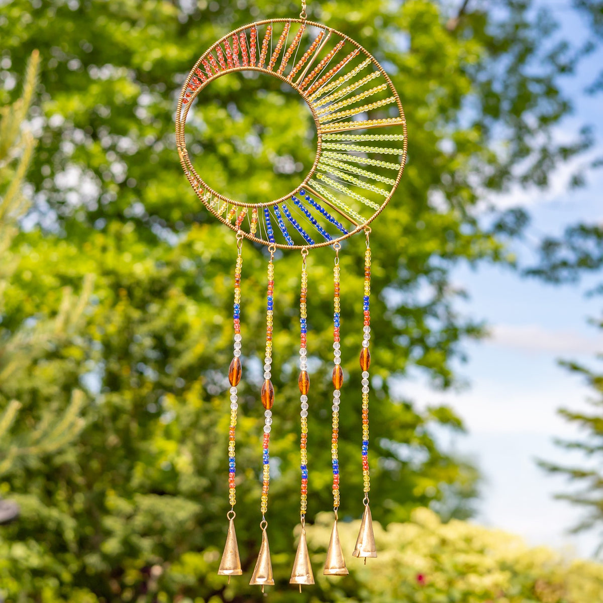 A circular gold-tone wind chime with colorful beadwork and hanging bell-shaped pieces against leafy trees.