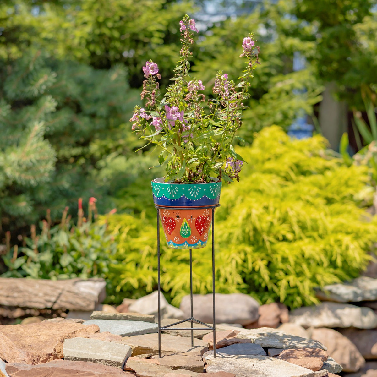 Flowerpot with a teal rim and orange floral design on a black iron stand among garden stones.