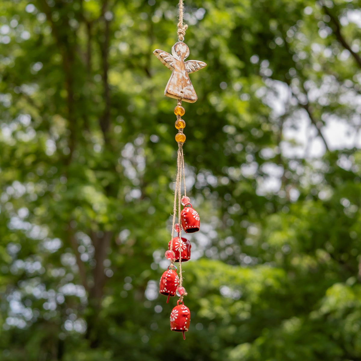 A tan angel wind chime with orange beads and red dangling bells hangs against leafy trees.