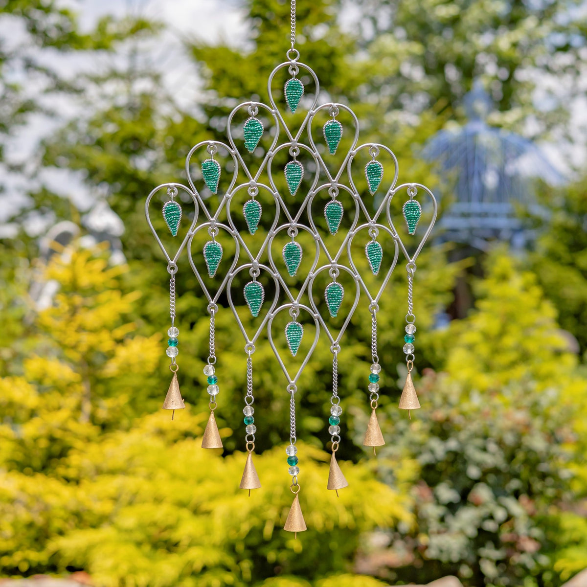 Silver-tone wind chime with teal glass accents and dangling bell-shaped pieces hanging among garden greenery.