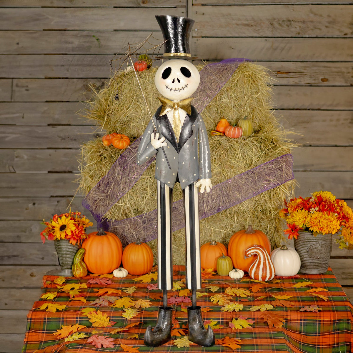 Full-length skeleton figure in a top hat and suit, holding a spiderweb umbrella amid autumn decorations.